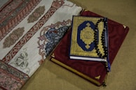A Quran with a blue and gold ornate cover rests on top of a red prayer rug adorned with intricate embroidery. Beside it, a section of an intricately designed fabric with floral and geometric patterns is visible.