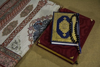 A Quran with a blue and gold ornate cover rests on top of a red prayer rug adorned with intricate embroidery. Beside it, a section of an intricately designed fabric with floral and geometric patterns is visible.