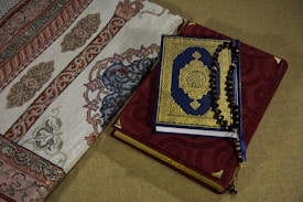 A Quran with a blue and gold ornate cover rests on top of a red prayer rug adorned with intricate embroidery. Beside it, a section of an intricately designed fabric with floral and geometric patterns is visible.