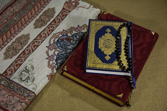 A Quran with a blue and gold ornate cover rests on top of a red prayer rug adorned with intricate embroidery. Beside it, a section of an intricately designed fabric with floral and geometric patterns is visible.