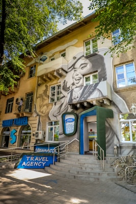 A travel agency is located in a multi-story building adorned with a large mural. The mural depicts a smiling woman's face with her hand raised as if waving. The building is surrounded by trees, and there is a sign indicating 'TRAVEL AGENCY' at the entrance. Several windows and architectural details are visible, as well as an entrance with a ramp and stairs.