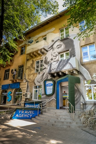 A travel agency is located in a multi-story building adorned with a large mural. The mural depicts a smiling woman's face with her hand raised as if waving. The building is surrounded by trees, and there is a sign indicating 'TRAVEL AGENCY' at the entrance. Several windows and architectural details are visible, as well as an entrance with a ramp and stairs.