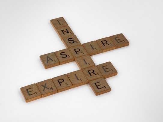 Wooden Scrabble tiles are arranged to form the words 'INSPIRE', 'ASPIRE', and 'EXPIRE' in a crossword style on a white background.