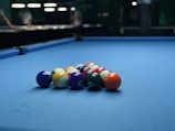 A set of billiard balls arranged in a triangular formation on a blue pool table. The background is slightly blurred, with dim lighting and some figures visible.