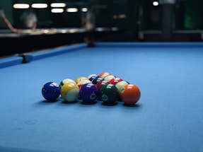 A set of billiard balls arranged in a triangular formation on a blue pool table. The background is slightly blurred, with dim lighting and some figures visible.