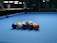 A set of billiard balls arranged in a triangular formation on a blue pool table. The background is slightly blurred, with dim lighting and some figures visible.