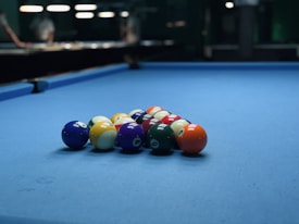 A set of billiard balls arranged in a triangular formation on a blue pool table. The background is slightly blurred, with dim lighting and some figures visible.
