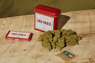 A red tin labeled 'Dad Grass' is positioned on a wooden surface alongside a rectangular box with similar branding. A pile of green, dried hemp or marijuana flower sits nearby, and a small brown packet labeled for humidity control is included.
