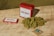 A red tin labeled 'Dad Grass' is positioned on a wooden surface alongside a rectangular box with similar branding. A pile of green, dried hemp or marijuana flower sits nearby, and a small brown packet labeled for humidity control is included.