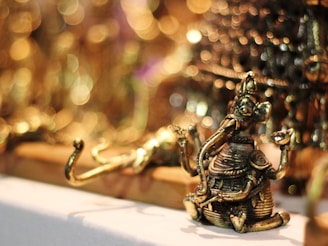 A small, intricately detailed brass figure of a deity with an elephant head, seated with one knee up and holding objects in its hands. The background features a soft focus with circular bokeh lights, giving a warm and luminous ambiance.