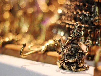A small, intricately detailed brass figure of a deity with an elephant head, seated with one knee up and holding objects in its hands. The background features a soft focus with circular bokeh lights, giving a warm and luminous ambiance.