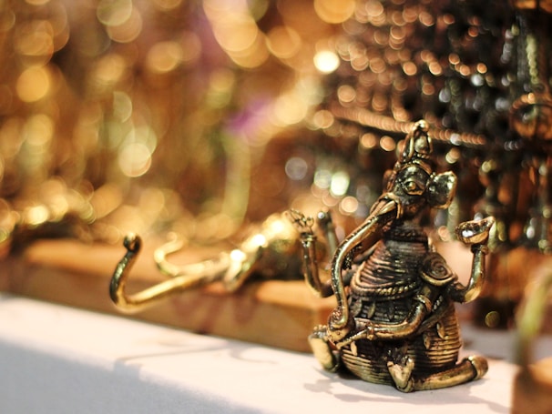 A small, intricately detailed brass figure of a deity with an elephant head, seated with one knee up and holding objects in its hands. The background features a soft focus with circular bokeh lights, giving a warm and luminous ambiance.