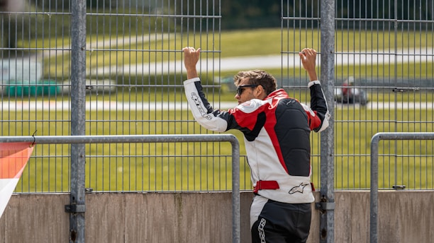 A person in a racing suit leans against a metal fence, looking out at a large open area with a road or track visible in the background. The individual is wearing sunglasses and has short hair, with the racing suit prominently displaying a red, black, and white color scheme.