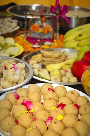A variety of traditional Indian sweets and fruits are presented on several plates. The focal point is a plate of round sweets garnished with rose petals. In the background, there are bananas, apple, and other dishes. An ornate decorated altar with vibrant orange marigold flowers and a purple ribbon is also visible.