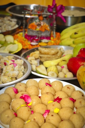 A variety of traditional Indian sweets and fruits are presented on several plates. The focal point is a plate of round sweets garnished with rose petals. In the background, there are bananas, apple, and other dishes. An ornate decorated altar with vibrant orange marigold flowers and a purple ribbon is also visible.