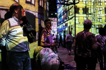 People are standing and walking in a dimly lit room with colorful digital projections on the walls, creating an immersive art experience. The projections depict a detailed building facade. The atmosphere appears lively with several individuals engaging with the artwork.