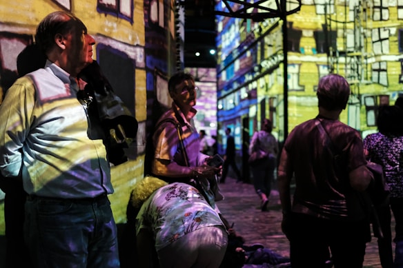 People are standing and walking in a dimly lit room with colorful digital projections on the walls, creating an immersive art experience. The projections depict a detailed building facade. The atmosphere appears lively with several individuals engaging with the artwork.