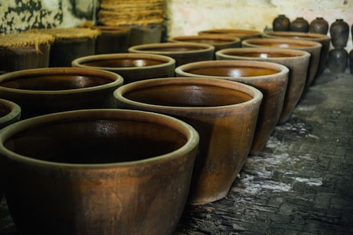 A row of large, rustic, brown ceramic pots are placed on a paved floor inside a dimly lit room. The pots are aligned with several others in the background. Some pottery items with a different shape and size are also visible in the background.