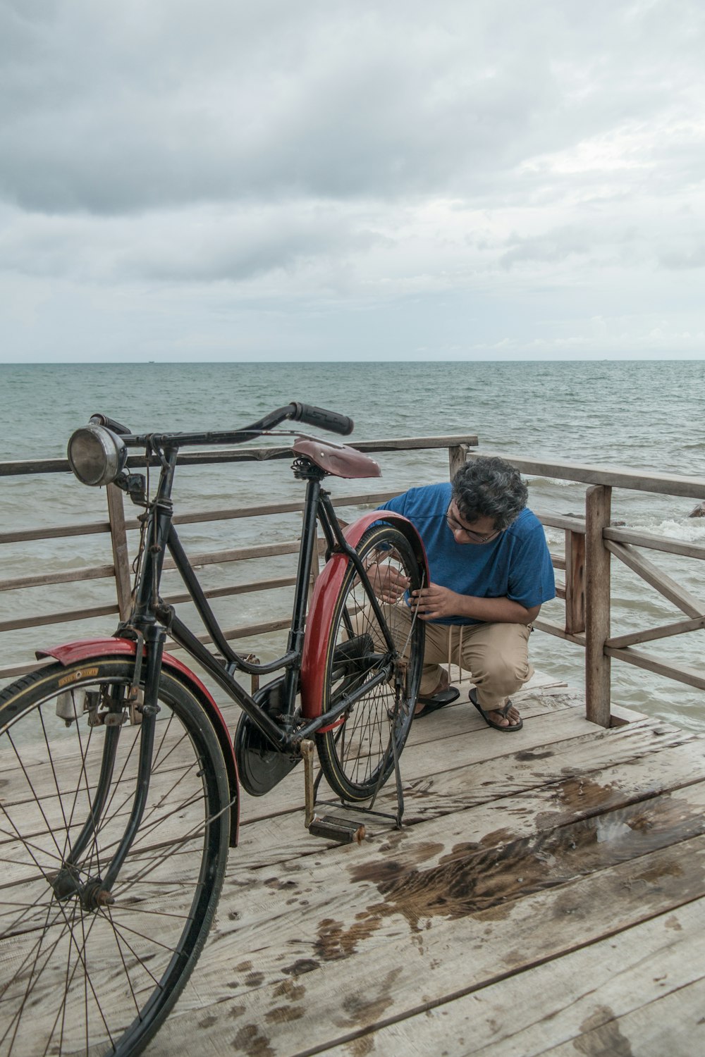 man in blue t-shirt sitting on brown wooden fence near body of water during daytime