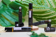 A variety of brown glass vials labeled 'Scentl.' are placed on a wooden surface surrounded by large green tropical leaves. Some vials are lying down, while others stand upright, and one appears to have an open cap.