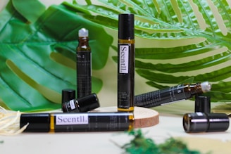 A variety of brown glass vials labeled 'Scentl.' are placed on a wooden surface surrounded by large green tropical leaves. Some vials are lying down, while others stand upright, and one appears to have an open cap.