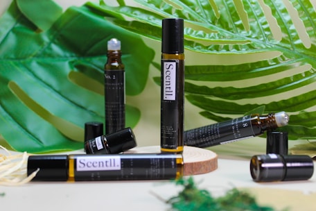 A variety of brown glass vials labeled 'Scentl.' are placed on a wooden surface surrounded by large green tropical leaves. Some vials are lying down, while others stand upright, and one appears to have an open cap.