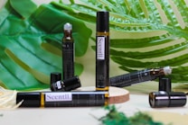 A variety of brown glass vials labeled 'Scentl.' are placed on a wooden surface surrounded by large green tropical leaves. Some vials are lying down, while others stand upright, and one appears to have an open cap.