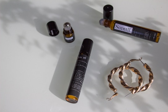 A flat lay of various lifestyle items arranged on a smooth surface. An open fragrance bottle with a rollerball sits with its cap next to it, another similar bottle on its side labeled 'Scentl’, and a pair of twisted hoop earrings made of a metallic material. Soft shadows from the items create a nuanced pattern on the surface.