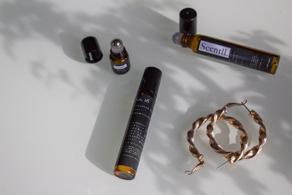 A flat lay of various lifestyle items arranged on a smooth surface. An open fragrance bottle with a rollerball sits with its cap next to it, another similar bottle on its side labeled 'Scentl’, and a pair of twisted hoop earrings made of a metallic material. Soft shadows from the items create a nuanced pattern on the surface.