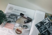 An open magazine featuring an article titled 'Downtime' with various images and a headline indicating a special focus on female voices. There is a close-up of a cup of coffee with frothy milk and cinnamon, a plant, an open book displaying its cover 'The White Album' by Joan Didion, a small cylindrical object resembling a perfume bottle, black and white photos, and a couple of rings.