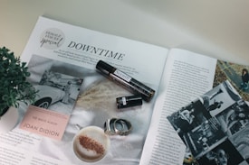 An open magazine featuring an article titled 'Downtime' with various images and a headline indicating a special focus on female voices. There is a close-up of a cup of coffee with frothy milk and cinnamon, a plant, an open book displaying its cover 'The White Album' by Joan Didion, a small cylindrical object resembling a perfume bottle, black and white photos, and a couple of rings.