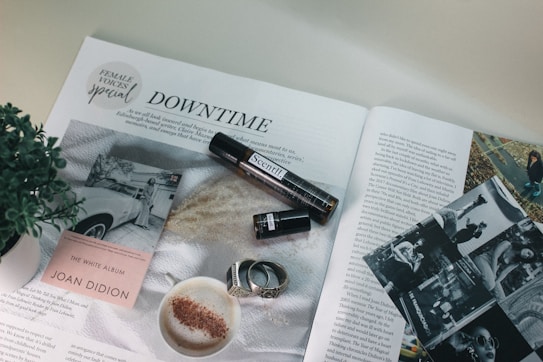An open magazine featuring an article titled 'Downtime' with various images and a headline indicating a special focus on female voices. There is a close-up of a cup of coffee with frothy milk and cinnamon, a plant, an open book displaying its cover 'The White Album' by Joan Didion, a small cylindrical object resembling a perfume bottle, black and white photos, and a couple of rings.
