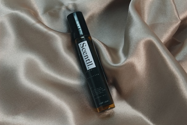 A small, dark glass bottle labeled 'Scentil' lies diagonally against a backdrop of smooth, beige satin fabric with flowing textures and soft folds. The label on the bottle is clear and elegant, adding a touch of luxury to the overall composition.