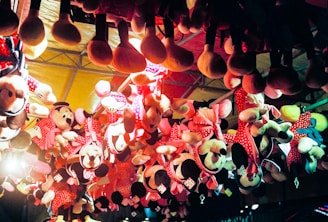 Several plush toys, mostly of a popular cartoon character, hang upside down from the ceiling as part of a carnival or amusement park prize display. The toys are predominantly dressed in red and pink polka-dotted outfits and have large round ears. The lighting gives the scene a warm, colorful glow, enhancing the festive atmosphere.