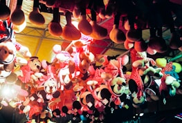 Several plush toys, mostly of a popular cartoon character, hang upside down from the ceiling as part of a carnival or amusement park prize display. The toys are predominantly dressed in red and pink polka-dotted outfits and have large round ears. The lighting gives the scene a warm, colorful glow, enhancing the festive atmosphere.