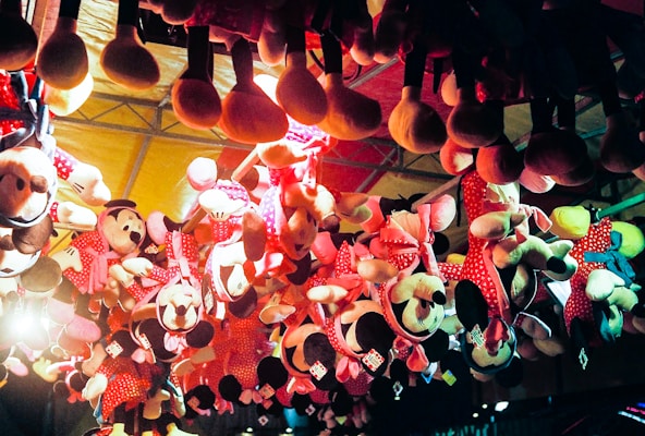 Several plush toys, mostly of a popular cartoon character, hang upside down from the ceiling as part of a carnival or amusement park prize display. The toys are predominantly dressed in red and pink polka-dotted outfits and have large round ears. The lighting gives the scene a warm, colorful glow, enhancing the festive atmosphere.