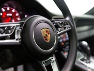 A close-up view of a car's steering wheel, prominently featuring the Porsche emblem. The interior of the car includes visible elements such as a digital dashboard with red illumination and a gear shift lever. The materials appear sleek and modern, with polished metal accents and a leather finish.