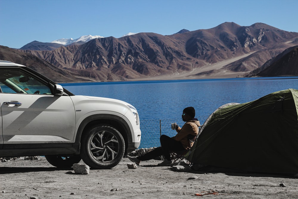 man in black jacket sitting on black camping chair near white car during daytime