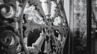 An intricate wrought iron design with a circular motif, characterized by floral and geometric patterns, against a blurred background. The ornamental metalwork is aged and slightly weathered, providing a sense of history and craftsmanship.
