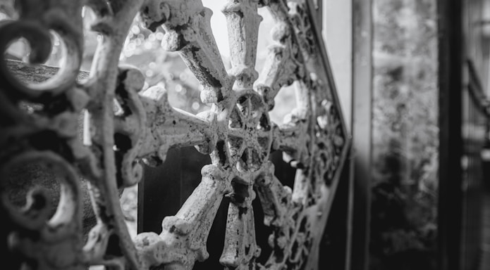 An intricate wrought iron design with a circular motif, characterized by floral and geometric patterns, against a blurred background. The ornamental metalwork is aged and slightly weathered, providing a sense of history and craftsmanship.
