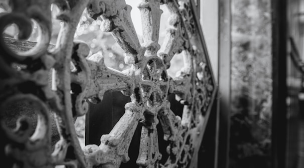 An intricate wrought iron design with a circular motif, characterized by floral and geometric patterns, against a blurred background. The ornamental metalwork is aged and slightly weathered, providing a sense of history and craftsmanship.