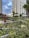 A modern urban garden with lush greenery, including various plants and small palm trees, is set against the backdrop of tall, contemporary apartment buildings. The sky is partly cloudy, creating a contrasting atmosphere between the natural elements and urban structures.