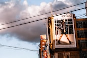 A large urban billboard displaying an advertisement featuring a person walking confidently in fashionable attire against a cityscape background. The scene is lit by warm, late afternoon sunlight, casting strong shadows. Overhead power lines and modern high-rise buildings add to the urban atmosphere of the image.