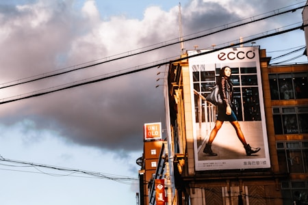 A large urban billboard displaying an advertisement featuring a person walking confidently in fashionable attire against a cityscape background. The scene is lit by warm, late afternoon sunlight, casting strong shadows. Overhead power lines and modern high-rise buildings add to the urban atmosphere of the image.
