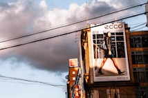 A large urban billboard displaying an advertisement featuring a person walking confidently in fashionable attire against a cityscape background. The scene is lit by warm, late afternoon sunlight, casting strong shadows. Overhead power lines and modern high-rise buildings add to the urban atmosphere of the image.