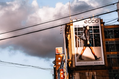 A large urban billboard displaying an advertisement featuring a person walking confidently in fashionable attire against a cityscape background. The scene is lit by warm, late afternoon sunlight, casting strong shadows. Overhead power lines and modern high-rise buildings add to the urban atmosphere of the image.