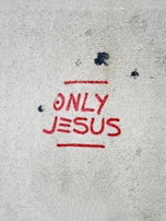 A concrete surface with the words 'ONLY JESUS' spray-painted in bold red letters, accompanied by a red underline. Several dark spots and stains are scattered across the surface, giving it a slightly weathered appearance.