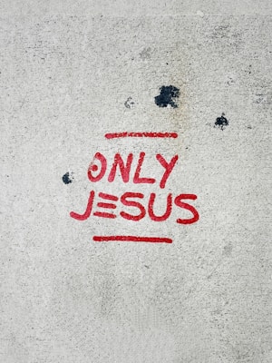 A concrete surface with the words 'ONLY JESUS' spray-painted in bold red letters, accompanied by a red underline. Several dark spots and stains are scattered across the surface, giving it a slightly weathered appearance.