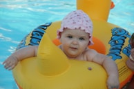 A baby with a pink floral-patterned hat is sitting in a yellow inflatable pool toy in the shape of a duck. The water in the pool is a clear blue, and the baby is smiling.