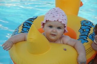 A baby with a pink floral-patterned hat is sitting in a yellow inflatable pool toy in the shape of a duck. The water in the pool is a clear blue, and the baby is smiling.